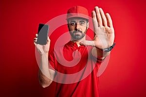 Young handsome delivery man with beard holding smartphone showing deliver food app with open hand doing stop sign with serious and