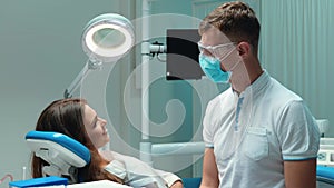 Young handsome dentist in medical blue gloves , mask and glasses talks to beautiful patient woman before starting