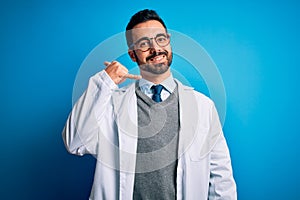 Young handsome doctor man with beard wearing coat and glasses over blue background smiling doing phone gesture with hand and