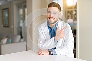 Young handsome doctor man at the clinic cheerful with a smile of face pointing with hand and finger up to the side with happy and