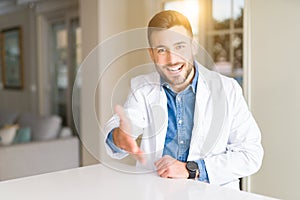 Young handsome doctor man at the clinic smiling friendly offering handshake as greeting and welcoming