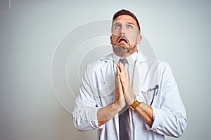 Young handsome doctor man wearing white profressional coat over  background begging and praying with hands together with