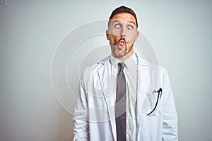 Young handsome doctor man wearing white profressional coat over isolated background making fish face with lips, crazy and comical