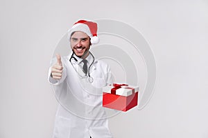 Young handsome doctor in white uniforme and Santa Claus hat standing in studio on white background smile and finger in camera photo