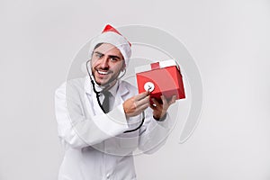 Young handsome doctor in white uniforme and Santa Claus hat standing in studio on white background smile and finger in camera photo