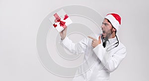 Young handsome doctor in white uniforme and Santa Claus hat standing in studio on white background smile and Shows a finger at the photo