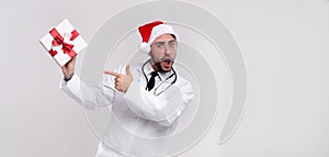 Young handsome doctor in white uniforme and Santa Claus hat standing in studio on white background smile and Shows a finger at the photo