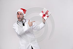 Young handsome doctor in white uniforme and Santa Claus hat standing in studio on white background smile and Shows a finger at the photo