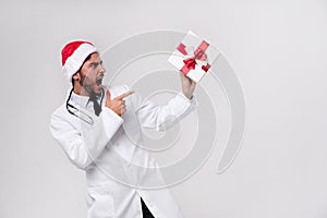 Young handsome doctor in white uniforme and Santa Claus hat standing in studio on white background smile and Shows a finger at the photo