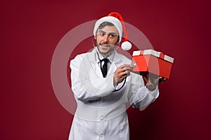 Young handsome doctor in white uniforme and Santa Claus hat standing in studio on red background smile and finger in camera photo