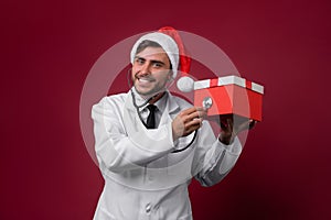 Young handsome doctor in white uniforme and Santa Claus hat standing in studio on red background smile and finger in camera photo