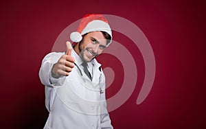 Young handsome doctor in white uniforme and Santa Claus hat standing in studio on red background smile and finger in camera photo