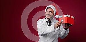 Young handsome doctor in white uniforme and Santa Claus hat standing in studio on red background smile and finger in camera photo