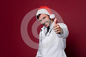 Young handsome doctor in white uniforme and Santa Claus hat standing in studio on red background smile and showing a thumbs up photo