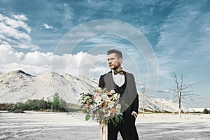 Young handsome elegant man in fashionable suit and bow tie is holding a big bouquet of flowers and looks to side at the