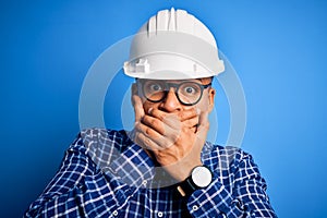 Young handsome engineer latin man wearing safety helmet over isolated blue background shocked covering mouth with hands for