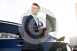 Young handsome European businessman at charging station, standing near electric car with laptop pc and looking away