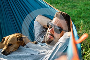 Young handsome European man in sunglasses is resting in hammock with his cute little dog