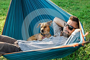 Young handsome European man in sunglasses is resting in hammock with his cute little dog