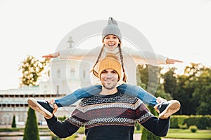 Young handsome father gives piggyback to his little smiling daughter, have fun together when have excursion. Small child looks at