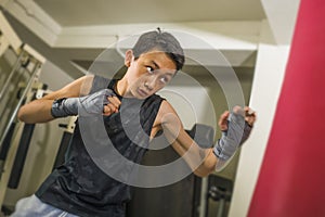 Young handsome and fierce 13 or 14 years old boy in wrist wraps doing fight workout punching heavy bag looking cool and badass at