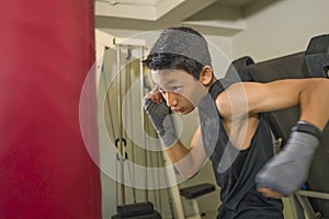 Young handsome and fierce 13 or 14 years teenager boy in wrist wraps doing fight workout punching heavy bag looking cool and