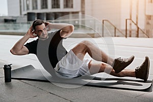 Young handsome fit brunette man in sportswear warming up before street workout doing crunches on yoga mat, healthy fit