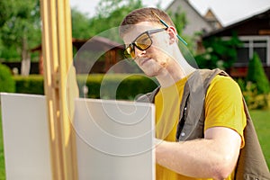 Young handsome guy artist painting picture on canvas outside in park, outdoors