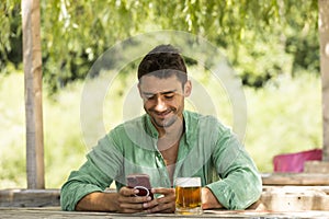 Young, handsome guy in the middle of summer sitting at a table with a beer in nature while looking at his phone smiling