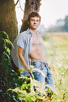 Young handsome guy with a naked torso with elastic belly muscles in an unbuttoned shirt posing on nature in a meadow near a t