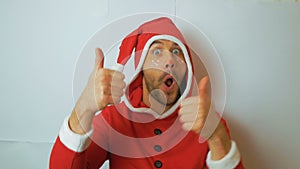 A young handsome guy in a red santaclaus carnival costume shakes his head and smiles broadly and shows a thumbs up. A