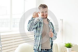 Young handsome guy shows the keys to his new apartment and showing thumbs up while standing in the living room. Mortgage