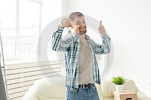 Young handsome guy shows the keys to his new apartment and showing thumbs up while standing in the living room. Mortgage