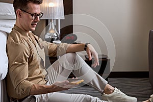 Young handsome guy sit on floor in hotel room, having rest, chatting with friends on mobile phone