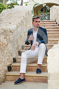 Young handsome guy in a suit and white trousers sits on the stairs