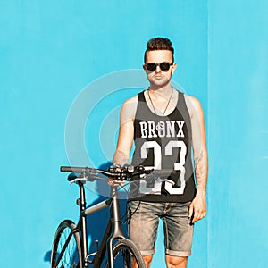 Young handsome guy in sunglasses, a black T-shirt and denim shorts riding a bicycle.on the background of a bright blue wall.