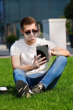 Young handsome guy with sunglasses using tablet and headphones, drinking coffee to go, sitting on grass and enjoying nice day.