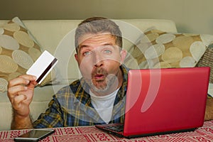Young handsome and happy man holding credit card sitting at home sofa couch using laptop computer online shopping smiling cheerful