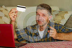 Young handsome and happy man holding credit card sitting at home sofa couch using laptop computer online shopping smiling cheerful