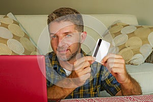 Young handsome and happy man holding credit card sitting at home sofa couch using laptop computer online shopping smiling cheerful