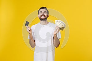 Young handsome happy smiling man holding banking card and cash in his hands isolated on yellow background.