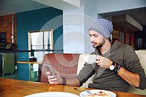 Young handsome hipster man with beard sitting in cafe using a mobile phone, holding cup of coffee.