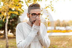 Young handsome hipster man in glasses and a white sweater is walking in a cold autumn park. Heats his hands with his breath.