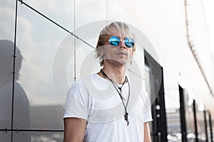 Young handsome hipster man in vintage t-shirt with amulets on a neck in stylish sunglasses with trendy hairstyle stands near the