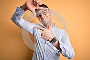 Young handsome hispanic business man wearing nerd glasses over yellow background smiling making frame with hands and fingers with