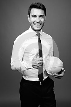 Young handsome Hispanic businessman with hardhat against gray background