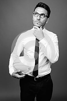 Young handsome Hispanic businessman with hardhat against gray background