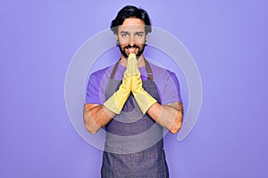 Young handsome hispanic clenaer man wearing housework apron and washing gloves praying with hands together asking for forgiveness