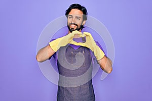 Young handsome hispanic clenaer man wearing housework apron and washing gloves smiling in love doing heart symbol shape with hands