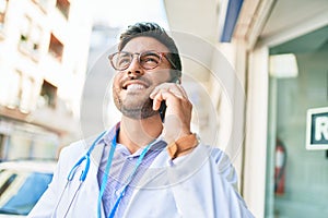 Young handsome hispanic doctor wearing stethoscope smiling happy Standing with smile on face talking on the smartphone at town